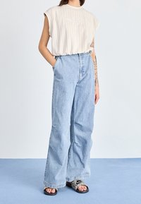 Kvinna iklädd en ärmlös beigefärgad randig topp, ljusblå vida jeans med händerna i fickorna och Nike-slides med ormskinnsmönster.