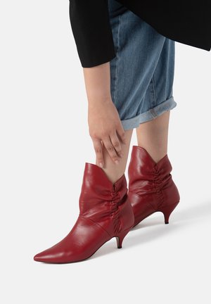 Stivaletti in pelle rossa con punta affilata, dettagli arricciati alla caviglia e tacco stiletto basso. Indossati con jeans arrotolati.