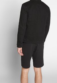 Veste en denim noir avec des accents côtelés sur les manches, associée à un short noir. Vue de l'arrière, mettant en valeur la texture et l'ajustement du matériau.