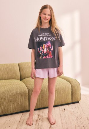 Girl with long blonde hair stands barefoot wearing a black K-pop Huntrx Demon Hunters T-shirt and pink shorts in a bright room with a green sofa.