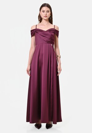 Donna con un abito lungo fino a terra in raso color bordeaux con spalle scoperte e spalline sottili, in piedi davanti a uno sfondo chiaro semplice.