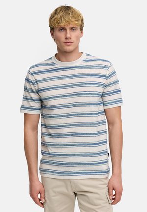 Blondyn ubrany w białą koszulkę z krótkim rękawem z poziomymi niebieskimi paskami i beżowe spodnie, stojący na jednolitym tle.