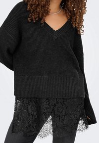 Femme portant un pull en maille noir à col en V superposé sur une jupe ou robe en dentelle fleurie noire, avec des cheveux bouclés et un collier en chaîne argentée.