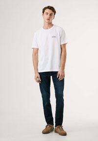 Hvit bomull t-skjorte med korte ermer som har en liten trykt logo på brystet, sammen med mørkeblå denimjeans og brune sko.
