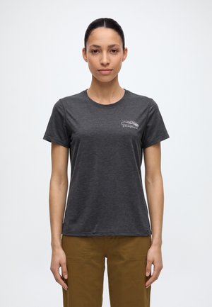 Une femme porte un tee-shirt Patagonia gris anthracite avec un petit logo sur la poitrine et un pantalon marron, sur un fond clair uni.