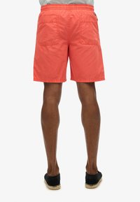 Shorts en coton corail avec une taille élastique, deux poches arrière et une coupe droite. Portés avec des espadrilles noires.