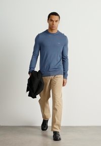Light blue knit sweater with a crew neck, paired with beige trousers and black shoes. Holding a black jacket in left hand.