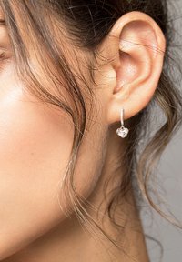 Silverfärgade hjärtformade örhängen med en polerad yta, hängande från en tunn ring, presenterade mot en texturerad hudbakgrund.