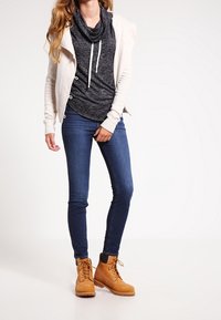 Beige kofta över en mörkgrå tröja med krage, tillsammans med blå slim fit jeans och tan snörskor med svart manschett.