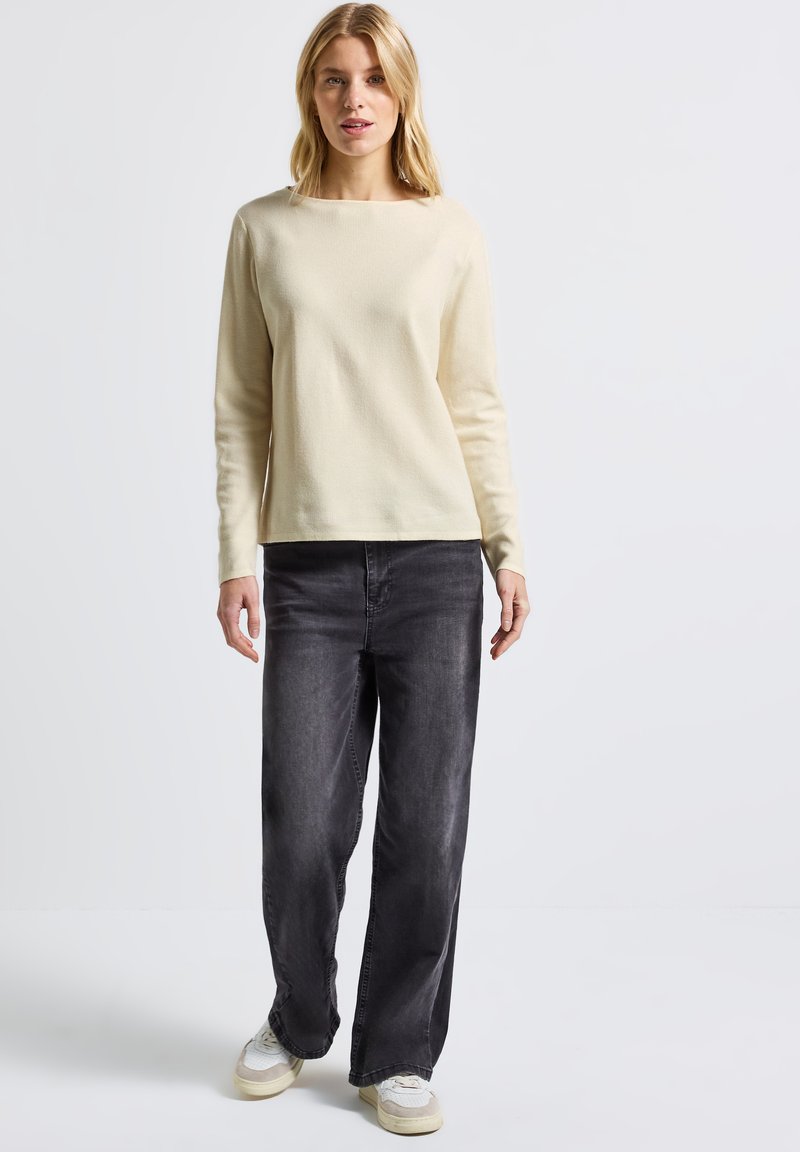 Maglione long sleeve beige chiaro con texture a coste, abbinato a jeans larghi a vita alta grigio scuro e scarpe da ginnastica bianche casual.