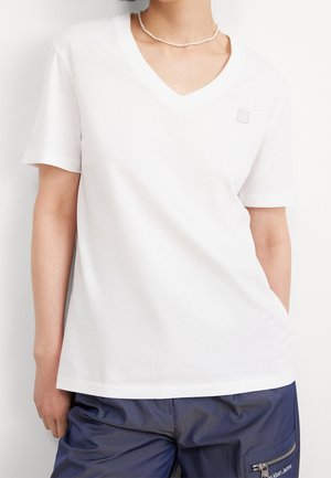 Person trägt ein schlichtes weißes V-Ausschnitt-T-Shirt mit kurzen Ärmeln und dunkelblaue Hosen mit Reißverschlusstasche, dazu eine einfache Perlenkette als Accessoire.