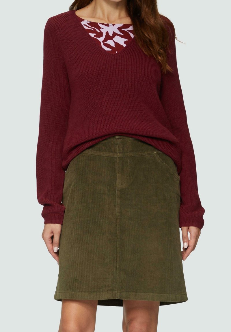 Femme portant un pull en maille bordeaux par-dessus une chemise à motifs et une jupe en velours côtelé vert olive avec des poches frontales.