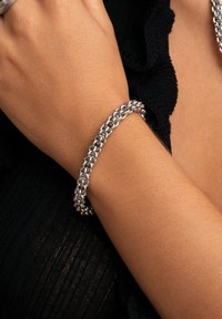 Bracelet en chaîne en argent avec des maillons ovales entrelacés et une finition polie, porté sur un poignet, sur un fond texturé sombre.