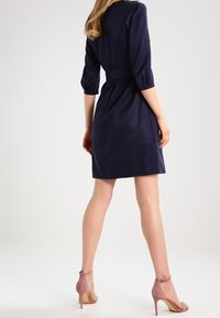 Námořnické modré šaty s úzkým pasem, tříčtvrtečními rukávy a sukní ve tvaru A ke kolenům, doplněné světlými růžovými lodičkami na vysokém podpatku.