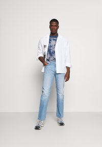 Camisa blanca de gran tamaño, camiseta azul con efecto tie-dye debajo, jeans rectos de color azul claro y zapatillas deportivas grises con acentos de colores.