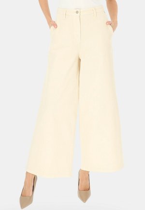 Kobieta ubrana w kremowe spodnie z wysokim stanem i szerokimi nogawkami z bocznymi kieszeniami, w połączeniu z beżowymi szpilkami w szpic, stojąca na jednolitym tle.