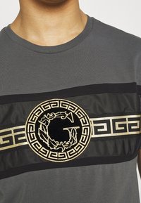 Šedé tričko s krátkým rukávem, které má černý a zlatý vyšitý emblém s bobkovými listy, doplněné kontrastními černými a zlatými vodorovnými pruhy.