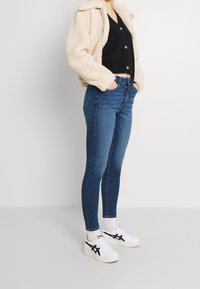 Giacca fluffy color crema sopra una maglietta nera, abbinata a jeans skinny blu e sneaker bianche con accenti neri.