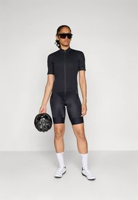 Crni biciklistički kombinezon s kratkim rukavima, patentnim zatvaračem sprijeda i kratke hlače s jastučićima. Model drži crnu kacigu, nosi velike tamne sunčane naočale i bijele čarape.