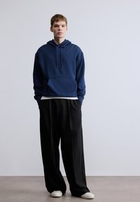 Hanorac bleumarin cu buzunar frontal și șnur, asortat cu pantaloni largi negri și pantofi albi slip-on. Textură netedă a țesăturii.