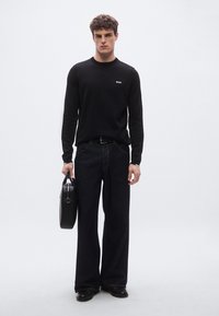 Musta pitkähihainen paita, jossa pieni valkoinen logo, yhdistetty leveälahkeisiin tummansinisin farkkuihin. Malli pitää kädessään mustaa jämäkkää käsilaukkua.