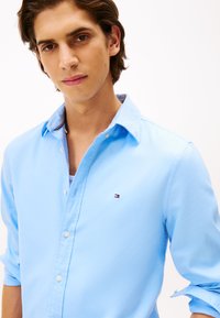 Hellblaues button-up Hemd aus Baumwolle mit einem kleinen Logo auf der Brust, mit Kragen, hochgeschlagenen Ärmeln und weißen Knöpfen.