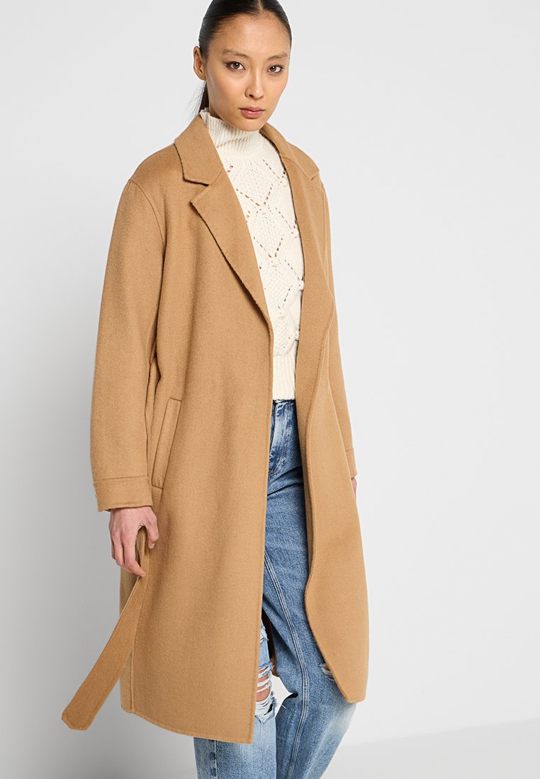 Płaszcz z mieszanki wełny w kolorze camel, długi, z klapami, przednimi kieszeniami i prostym krojem; noszony na kremowym swetrze w warkocze i zniszczonych dżinsach.