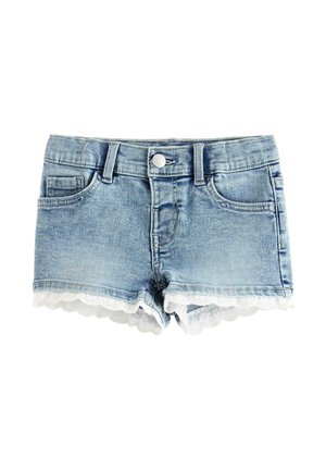 Ung pige med langt blondt hår iført en hvid broderet top, denimshorts med blondekant og blomstrede sløjfesko, står og smiler.