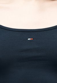 Κοντινό πλάνο σκούρου ναυτικού μπλουζιού με στρογγυλή λαιμόκοψη και μικρό ορθογώνιο λογότυπο σε λευκό και κόκκινο, τοποθετημένο στο κέντρο κάτω από τη λαιμόκοψη.