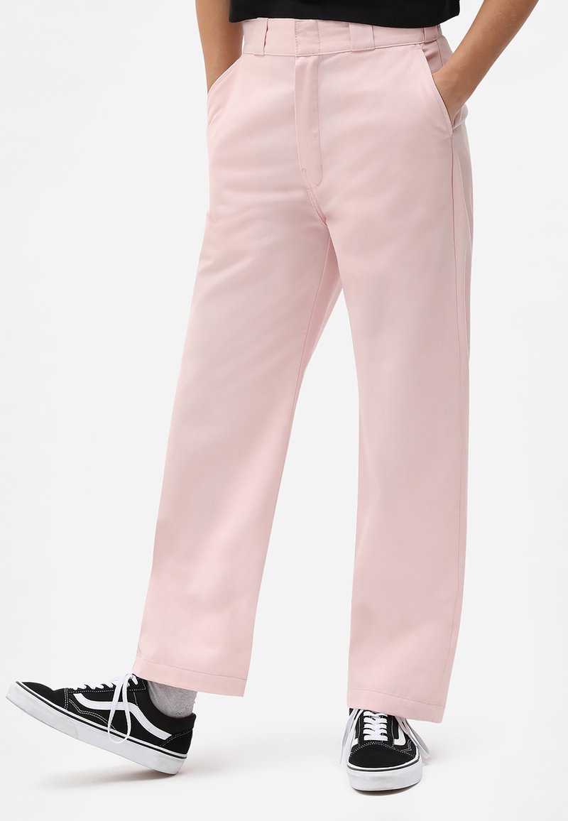 Personne portant un pantalon droit rose clair, les mains dans les poches et des baskets noires et blanches, debout sur un fond blanc.