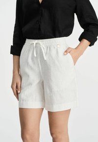 Białe lniane szorty z elastycznym paskiem, bocznymi kieszeniami i podwiniętym dołem. Stylizowane z czarną koszulą zapinaną na guziki. Miękka tekstura.