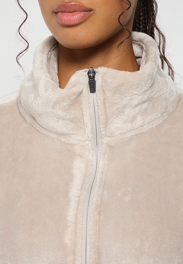 Beige Fleecejacke mit hohem Kragen, ausgestattet mit einem Frontreißverschluss und weichem Material. Sichtbare Nähte entlang der Kanten und des Reißverschlusses.