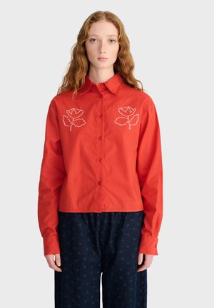 Jeune femme aux longs cheveux roux bouclés porte une chemise rouge boutonnée avec des motifs de feuilles blanches sur la poitrine et un pantalon ample sombre à motifs.
