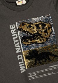 Harmaa t-paita, jossa on paljeteista koostuva dinosaurusten fossiilien grafiikka ja valkoinen teksti "WILD NATURE". Kuvan alla informatiivista tekstiä.