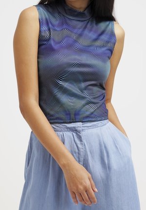 Vrouw draagt een mouwloze top met een hoge hals met blauwe en zwarte golvende lijnen en lichtblauwe, hoge taillebroek met zakken.