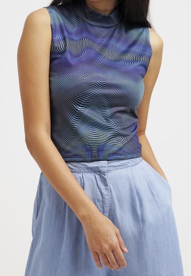 Femme portant un haut sans manches à col montant avec des lignes ondulées bleues et noires et un pantalon taille haute bleu clair avec des poches.