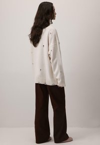 Przestronna kremowa sweter z nieregularnymi otworami, ściągacz u dołu, zestawiona z luźnymi ciemno-brązowymi spodniami. Miękki materiał, casualowy design.