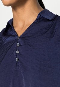 Camisa azul-marinho com um acabamento texturizado, apresentando um colarinho e cinco botões na parte da frente. O tecido parece leve e ligeiramente amarrotado.