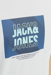 Valge puuvillane T-särk, millel on sinine ristkülikukujuline graafika diagonaalsete triipudega. Tekst "JACK&JONES" on kirjutatud paksude tähtedega, allpool on lisaks väiksem tekst.