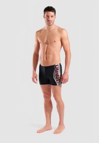 Czarne kąpielówki z czerwono-białym logo. Projekt obejmuje teksturowany materiał, dopasowany krój oraz długość do połowy uda.