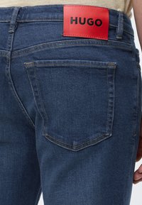 Poche arrière d'un jean en denim bleu foncé avec couture visible et étiquette rouge affichant "HUGO" en lettres noires sur la ceinture.