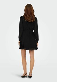 Czarna sukienka z szyfonu z długimi rękawami, paskiem w talii i rozkloszowanym dołem; noszona z czarnymi obcasami typu slingback.