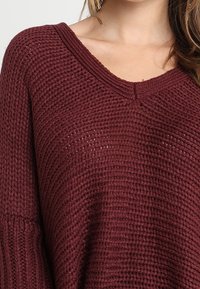 Burgundy stickad tröja med V-hals, med ett strukturerat stickmönster och sänkta axlar. Mjuk och mysig material.