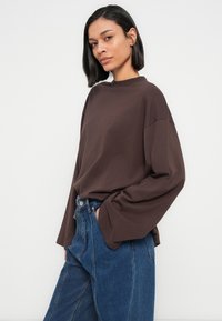 Καφέ μακρυμάνικη μπλούζα από απαλό ύφασμα, με ψηλό λαιμό και άνετη εφαρμογή, συνδυασμένη με ψηλόμεσο μπλε τζιν.