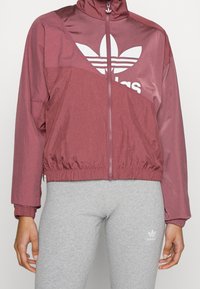 Vínová bunda na zip s límečkem a pružnými manžetami, s velkým bílým logem Adidas. Kombinováno se šedými přiléhavými legínami.