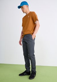 Braunes Kurzarm-T-Shirt mit weißem Logo, graue Hose und schwarze Schuhe. Eine blaue Kappe rundet den Look ab, während er auf grünem Boden steht.