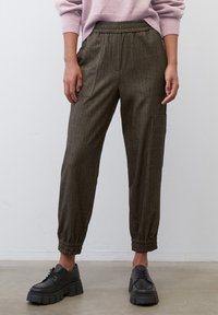 Bruine, gestreepte, taps toelopende broeken met een elastische tailleband en zijzakken, gecombineerd met zwarte chunky schoenen, op een gladde ondergrond.