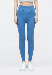 Kék leggings magas derékrésszel, rugalmas anyagból készült. Bal csípőjén kis fehér logó található, karcsú, testhezálló kialakítással.