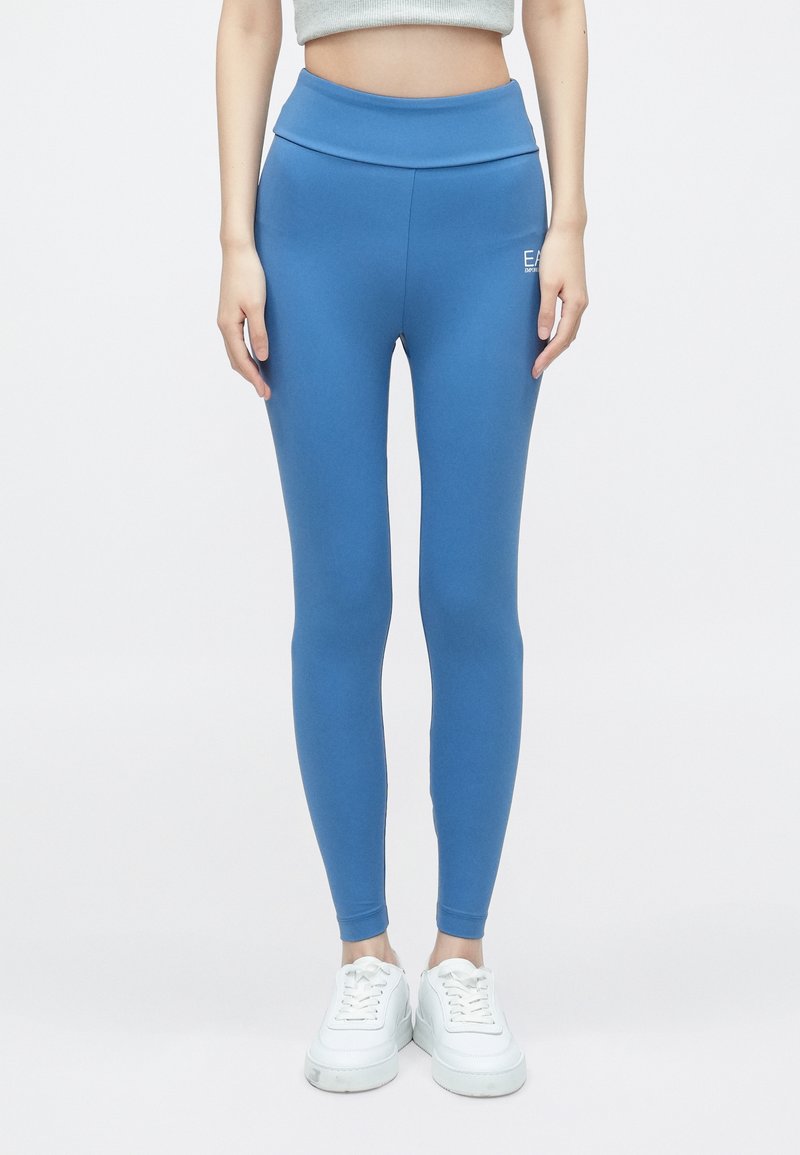 Kék leggings magas derékrésszel, rugalmas anyagból készült. Bal csípőjén kis fehér logó található, karcsú, testhezálló kialakítással.