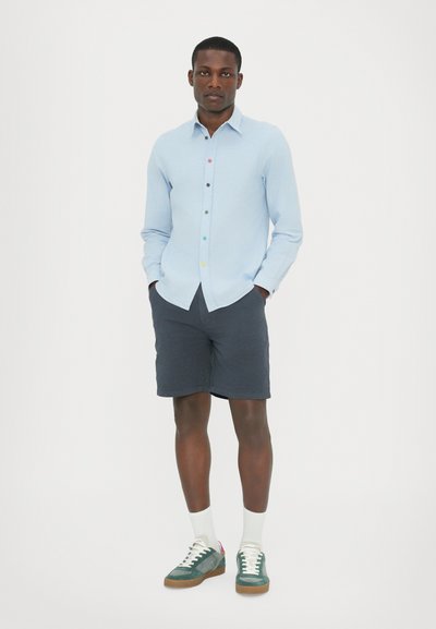 Ljust blå knappad skjorta med flerfärgade knappar, tillsammans med mörka shorts och gröna sneakers. Skjortan har en klassisk krage och lång ärm.